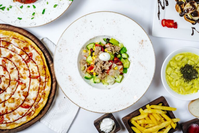 A colorful salad with vegetables and tuna, surrounded by pizza, fries, and dipping sauce.